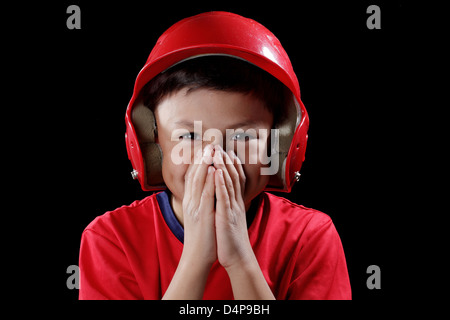 Sorridente giovane ragazzo copre la bocca con le mani - con il baseball rosso casco su sfondo nero - drammatico lo stile di illuminazione Foto Stock