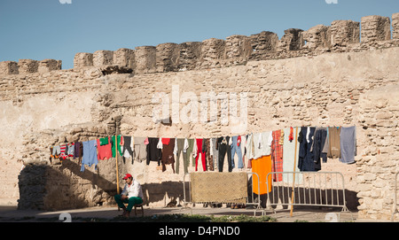 Un uomo seduto accanto alla biancheria stesa ad asciugare. Merlata cinta bastionata di Essaouira dietro. Foto Stock