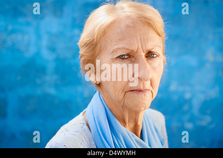Personale Senior ritratto, triste felice donna bionda in blu vestiti guardando la telecamera contro la parete blu Foto Stock