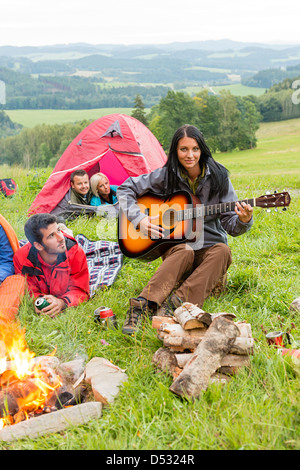 Giovane donna a suonare la chitarra accanto a camping amici giacente in tende Foto Stock