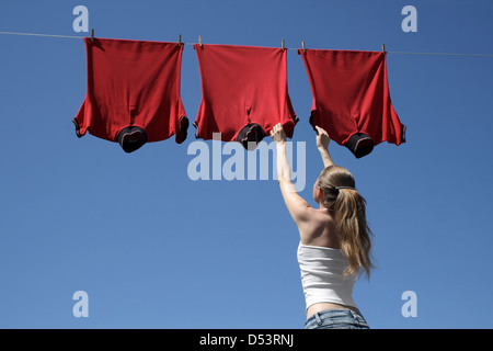 Giovane donna di raggiungere red corporate t-shirts appesi per asciugare in una brezza estiva su un vestiti-linea. Foto Stock