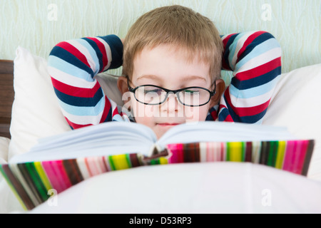 Poco carino boy in bicchieri libro di lettura a letto Foto Stock