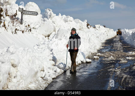 Un camminatore, Janet Lees, si trova all'incrocio tra la strada costiera Cronk-y-Voddy e Peel sull'Isola di Man, mentre una pesante scavatrice libera un sentiero attraverso la neve profonda sullo sfondo. Foto Stock