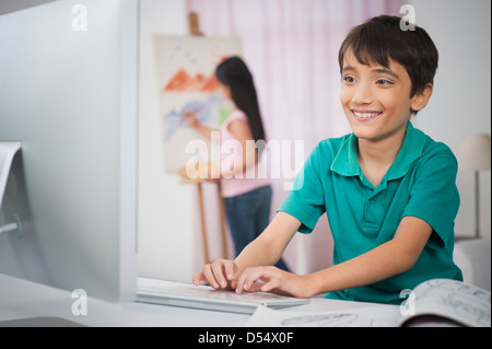 Boy utilizzando un PC desktop al tavolo da studio con sua sorella dipinto su tela di artisti Foto Stock