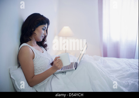 Donna seduta sul letto mediante un computer portatile mentre si beve il caffè Foto Stock