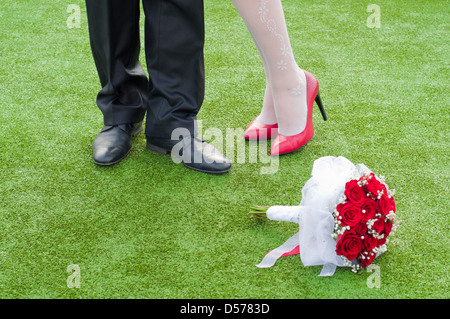 Le gambe della sposa e il fidanzato sull'erba verde con matrimonio mazzo rosso Foto Stock