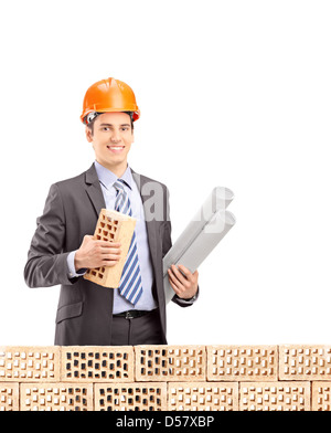 Giovane ingegnere maschio tenendo un mattone e un blueprint dietro un muro di mattoni isolati su sfondo bianco Foto Stock