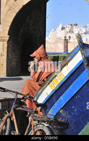 Uomo marocchino che indossa un Djellaba i cappotti tradizionali di tipo poncho invernale con cappuccio, seduto accanto alla barca vicino Bab El Marsa gate Essaouira, Marocco Foto Stock