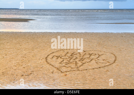 Felice Festa della Mamma scritta nel cuore di sabbia su una spiaggia di colore giallo, con un calmo mare blu in background Foto Stock