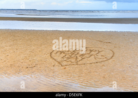 Felice Festa della Mamma scritta nel cuore di sabbia su una spiaggia di colore giallo, con un calmo mare blu in background Foto Stock