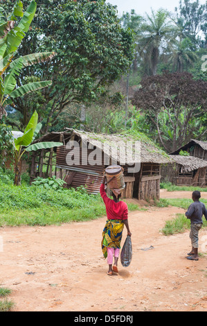 Donna africana a camminare sulla strada sterrata che porta la benna sulla testa Foto Stock