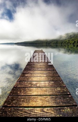 Il molo che si allunga oltre il lago calmo con nebbia di salita sulle colline, Rotorua, Nuova Zelanda Foto Stock