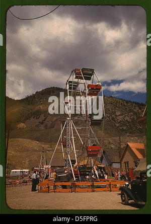 Questa immagine della fiera della contea di Delta del 1940 in Colorado mostra la scena del carnevale con una ruota panoramica e giostre in fiera. Cattura una vivace atmosfera rurale con veicoli e nuvole sullo sfondo, che riflettono la cultura rurale americana della metà del XX secolo e le attività ricreative. Foto Stock