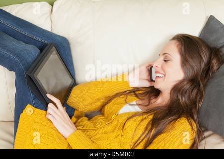 Donna sdraiata sul divano con tavoletta digitale e parlare attraverso il telefono cellulare Foto Stock