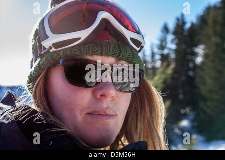 Giovane donna che guarda verso la fotocamera indossando occhiali da sole e un cappello di sci con occhiali protettivi sul suo capo. Foto Stock