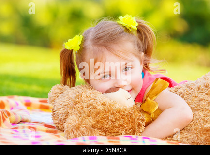 Carino sorridente bambina abbracciando soft bear toy, dolce kid avendo divertimento all'aperto in day care, che stabilisce su erba verde in primavera Foto Stock