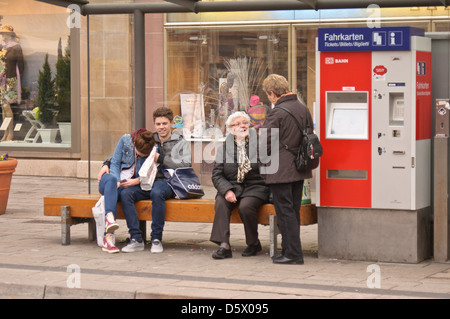 Giovani e vecchi. Adolescente matura un donne anziane in attesa ad una fermata del bus, stazione. Moderne Deutsche Bahn ticket machine - Heilbronn Germania Foto Stock