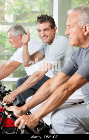 Felice l'uomo senior in palestra in bici tenendo pollice in alto Foto Stock