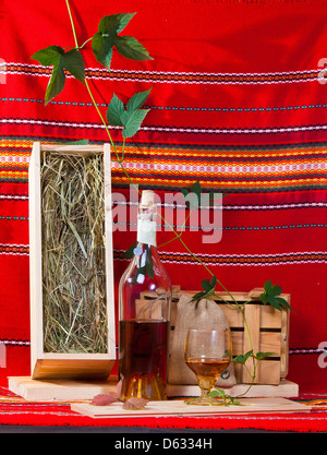 Still-life con una bottiglia di brandy, un vetro, scatole di legno e grapevine sulla stoffa rossa sullo sfondo Foto Stock