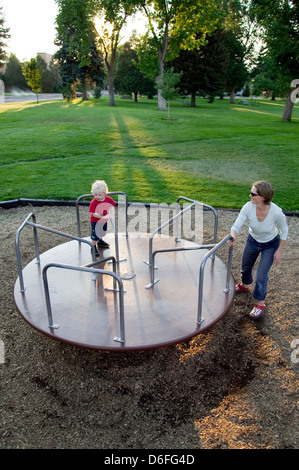 Giovane madre la filatura i suoi capelli biondi ragazzo su una merry-go-round su un parco giochi. Foto Stock