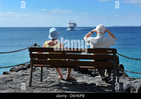 Una coppia di anziani seduti su una panchina a Playa Blanca, Lanzarote, guardando l'arrivo dell'Armas traghetto, Volcan De Tindaya. Foto Stock