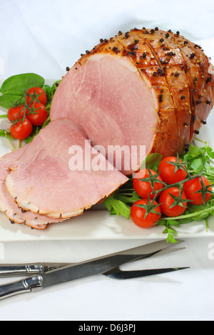 Il prosciutto cotto con chiodi di garofano e i pomodori ciliegia e carving coltello e forchetta su un quadrato bianco piastra su una tovaglia bianca Foto Stock