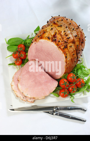 Il prosciutto cotto con chiodi di garofano e i pomodori ciliegia e carving coltello e forchetta su un quadrato bianco piastra su una tovaglia bianca - vista superiore Foto Stock