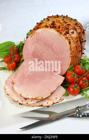 Il prosciutto cotto con chiodi di garofano e i pomodori ciliegia e carving coltello e forchetta su un quadrato bianco piastra su una tovaglia bianca Foto Stock