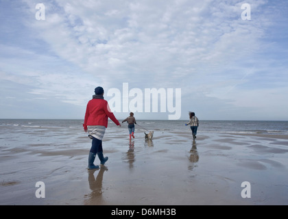 Tre donne camminando due cani su una spiaggia, spostandosi verso il mare. La donna in primo piano nella Red coat. Blue sky. Foto Stock