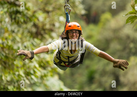 Due donne corrono in zipline fianco a fianco attraverso una fitta foresta verde. Entrambi indossano elmetti, guanti e imbracature arancioni collegati a un cavo. La donna davanti sorride con le braccia tese Foto Stock