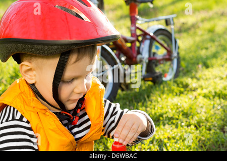 Little Boy fuori seduta in bicicletta sul prato verde di bere acqua in bottiglia con la sua moto parcheggiate a fianco e che indossa un casco di sicurezza Foto Stock