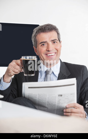 Felice manager seduto con giornali e caffè alla sua scrivania in ufficio Foto Stock