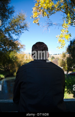 Vista posteriore di un uomo nel profondo del pensiero, appoggiato su di un ponte in un ambiente rurale. Foto Stock