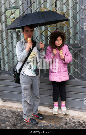 Leon e Maria a mangiare il gelato e riparo dalla pioggia sotto un ombrello a buon mercato in Piazza Navona, Roma, Italia Foto Stock
