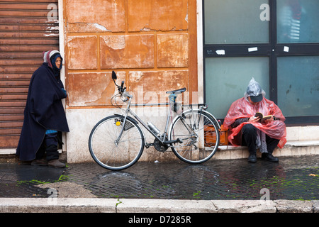 Senzatetto donna e un uomo che legge un libro sotto la pioggia sono separati da una bicicletta Foto Stock