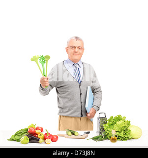 Gentiluomo maturo in posa con cucchiai e un libro di cucina durante la preparazione di un insalata isolati su sfondo bianco Foto Stock