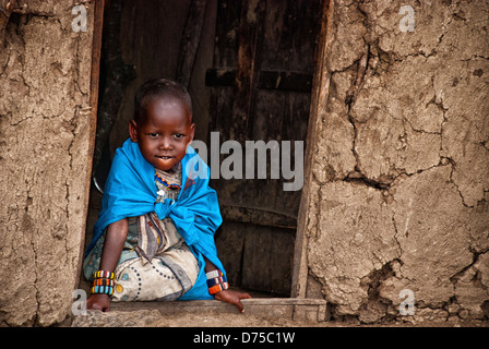 Masai Bambino, indossando abiti tradizionali, seduto alla porta di una capanna di fango in un villaggio Masai Mara, Kenya, Africa Foto Stock