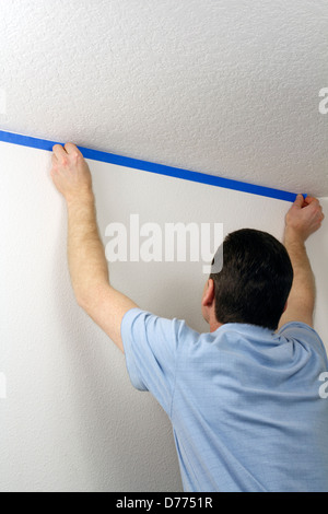 Uomo con nastro di pittori. Adulto maschio blu di pressatura pittore del nastro su una parete bianca sotto un soffitto al fine di proteggere la parete durante la verniciatura di soffitto. Foto Stock