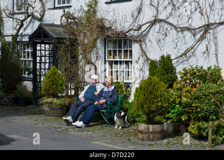 Coppia di anziani seduti su un banco di lavoro, con il cane, Hawkshead, Parco Nazionale del Distretto dei Laghi, Cumbria, England Regno Unito Foto Stock