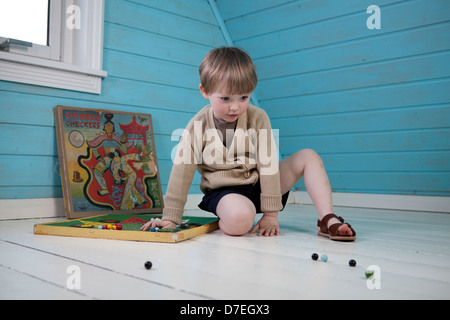 Un ragazzino in bei vestiti e sandali sta giocando da solo con il vecchio gioco di bordo in azzurro e bianco loft. Foto Stock