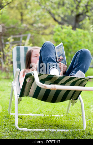 Ragazza adolescente sdraiato su un lettino prendisole e leggere un libro in giardino. Foto Stock