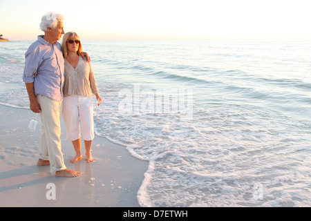 A piedi nudi coppia senior passeggiando lungo la spiaggia godendosi il tramonto Foto Stock