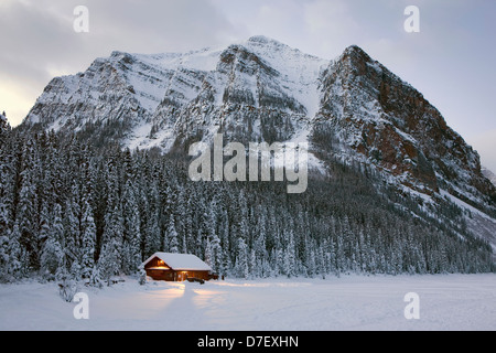 Una coperta di neve log cabin su lakeshore accesa fino al crepuscolo e circondato da coperta di neve alberi sempreverdi e montagne;il Lago Louise Foto Stock