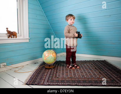 Little Boy tenendo il binocolo nelle sue mani in piedi accanto ad un globo luminoso che mostra e scolpito in legno elefante nella camera loft Foto Stock