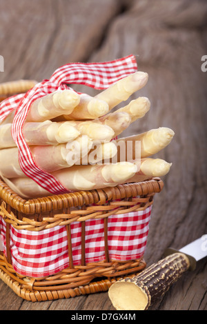 Asparagi bianchi legate con un nastro di tessuto in un piccolo cesto su una tavola di legno accanto a un coltello con impugnatura in corno di bue Foto Stock