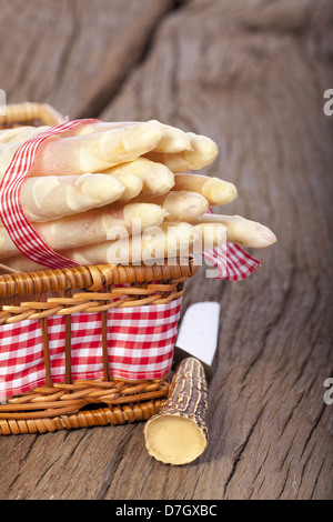 Asparagi bianchi legate con un nastro di tessuto in un piccolo cesto su una tavola di legno accanto a un coltello con impugnatura in corno di bue Foto Stock