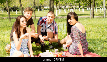 Amici dell'università avente un picnic nel parco con due coppie attraente seduti insieme su un tappeto gustando un pasto sano Foto Stock