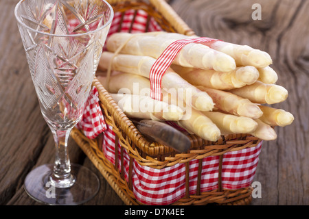 Asparagi bianchi legate con un nastro di tessuto in un piccolo cesto su una tavola di legno accanto a un bicchiere di vino Foto Stock