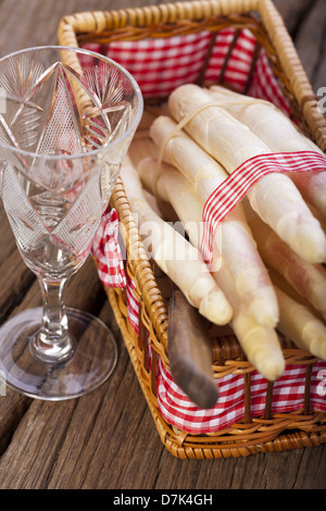 Asparagi bianchi legate con un nastro di tessuto in un piccolo cesto su una tavola di legno accanto a un bicchiere di vino Foto Stock