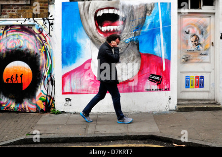 Un uomo a parlare su un telefono mobile, passeggiate passato un muro coperto di graffiti. Brick Lane, London, Regno Unito Foto Stock